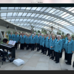 The Drybrook and district ladies choir stand together in a glass roofed building, they are wearing Blue blazers and black trousers